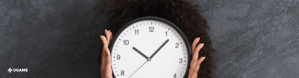 Esta imagem mostra uma pessoa segurando um relógio grande em frente ao rosto, cobrindo-o completamente. O relógio é redondo, com um fundo branco e números pretos, mostrando a hora aproximadamente 10:10. O fundo da imagem é cinza escuro e texturizado, e há um logotipo no canto inferior esquerdo com o texto "UGAME".