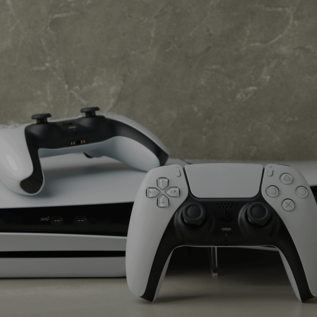 Um console de videogame moderno, acompanhado por dois controles, está sobre uma superfície neutra com um fundo de parede texturizada em tom cinza. O design do console e dos controles é elegante, predominando as cores branco e preto, com linhas suaves e detalhes minimalistas. A disposição dos controles, um em pé na frente e outro deitado sobre o console, destaca o equipamento e enfatiza o estilo sofisticado do dispositivo.